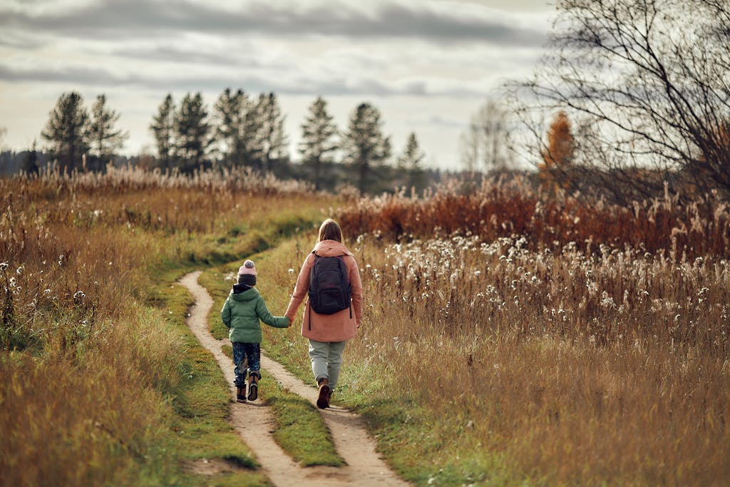 Back view of girl with backpack and little boy in hat with pompom walking by the hand along footpath through green field under cloudy sky