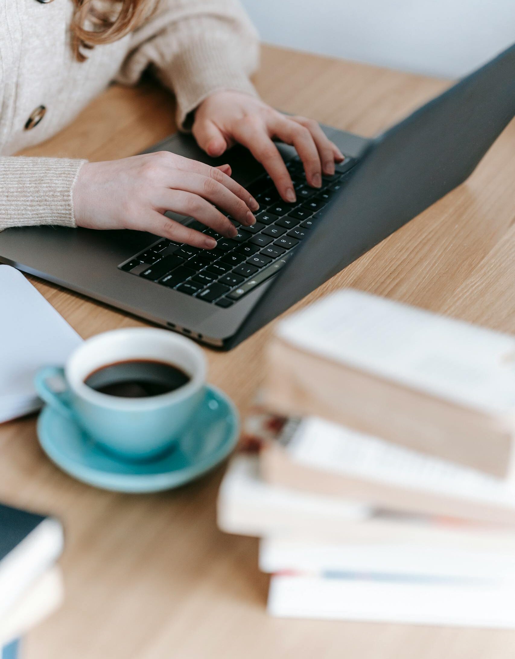 Crop anonymous female freelancer surfing netbook while sitting at wooden table with cup of hot coffee and books in room