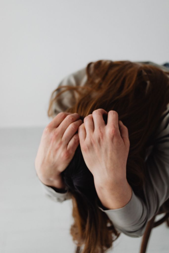 A woman with long hair holding her head, conveying stress and emotion against a neutral gray background.