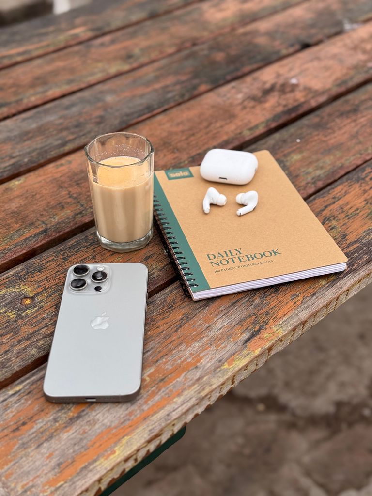 Outdoor scene with smartphone, earbuds, notebook and beverage on rustic table.
